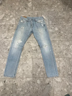 Ljusblå slitna jeans, storlek 34 - Säljer ett par ljusblå jeans med slitningar och distressed detaljer. Klassisk femficksmodell med raka ben och knappgylf. Jeansen är tillverkade i denim och har en avslappnad vibe som passar perfekt till streetwear.