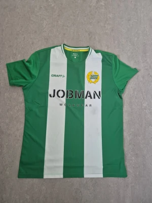 Grönvit Hammarby fotbollströja Craft - Säljer en grön och vit randig Hammarby fotbollströja från Craft. Tröjan har klubbmärke på bröstet, JOBMAN-tryck och klassisk rund hals med vit och grön kant. Tillverkad i polyester, perfekt för fotbollsträning eller match.