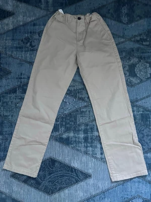 Beiga chinos med resårmidja - Snygga beiga chinos med rak passform och resår i midjan. Byxorna har klassisk design med knapp och dragkedja framtill samt fickor bak. Perfekta för en avslappnad men stilren look.