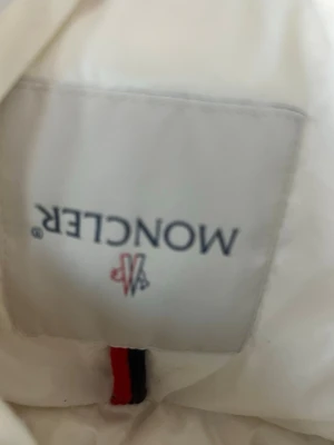 Vit dunjacka från Moncler - Snygg vit dunjacka från Moncler med klassisk logotyp på ärmen och detaljer i svart och rött. Jackan har en glansig finish och tryckknappar på fickorna. Perfekt för kalla vår och sommardagar och ger en clean, exklusiv look.
