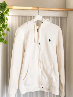Vit zip-hoodie från Polo Ralph Lauren - Snygg vit hoodie med dragkedja från Polo Ralph Lauren. Klassisk design med huva, dragsko och två fickor framtill. Ikoniska loggan broderad på bröstet. Perfekt för en clean och stilren look.
