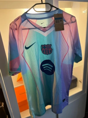 Nike FC Barcelona Dri-Fit matchtröja - Säljer en FC Barcelona matchtröja från Nike i Dri-Fit material. Tröjan har ett färgstarkt mönster i pastellfärger som blått, rosa och lila, med klubbmärke, Nike-logga och Spotify sponsortryck på bröstet. Perfekt för dig som älskar fotboll och vill sticka ut på planen.