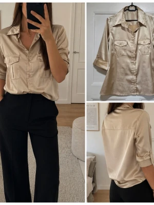 Champagnefärgad satinskjorta Savannah - Snygg champagnefärgad skjorta från Savannah i glansigt satinmaterial. Skjortan har trekvartsärmar, två bröstfickor med lock och knappar framtill. Perfekt för dig som gillar en elegant och trendig look.