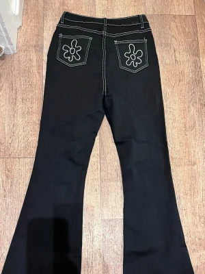 Svarta utsvängda jeans med kontrastsömmar🌸 - Oanvända Svarta jeans med utsvängda ben och vita kontrastsömmar. Bakfickorna har broderade blommönster som ger en cool retrovibe. Klassisk femficksmodell med knapp och dragkedja framtill. Perfekta för dig som gillar en unik och trendig stil.🧚🏻