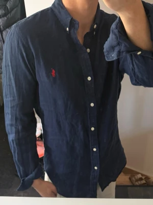 Marinblå linneskjorta från Ralph Lauren - Klassisk marinblå linneskjorta från Ralph Lauren med vit knappstängning och röd broderad logga på bröstet. Skjortan har lång ärm, button down-krage och är tillverkad i 100% linne. Perfekt för en stilren och avslappnad look.