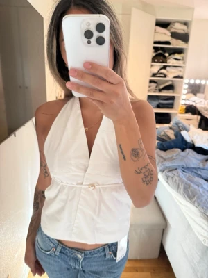 Vit halterneck topp med bälte och detalj  - Snygg vit halterneck topp med djup v-ringning och ett litet bälte med detalj framtill i midjan. Toppen är ärmlös och har en croppad passform som passar perfekt till jeans eller kjol. Tillverkad i ett mjukt bomullsmaterial.