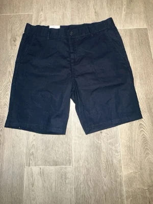 Svarta chinosshorts från C&A - Stilrena svarta chinosshorts från C&A med klassisk passform. Shortsen har sidofickor, bälteshällor och två bakfickor med knapp. Perfekta för en avslappnad och snygg look. Materialet är bomull för en skön känsla.