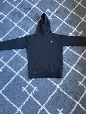 Svart hoodie från Lyle & Scott - Svart hoodie från Lyle & Scott med klassisk huva och känguruficka framtill. Liten gul logga på bröstet ger en clean och stilren look. Perfekt för dig som gillar enkel och snygg streetwear. Mjuk bomullskänsla och långa ärmar.