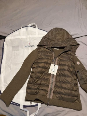 Brun dunväst med huva från Moncler - Snygg brun dunväst från Moncler med stickade ärmar och huva. Västen har dragkedja framtill, Moncler-logga på ärmen och ribbade muddar. Perfekt för lager-på-lager och streetstyle. Kommer med original Moncler-fodral.
