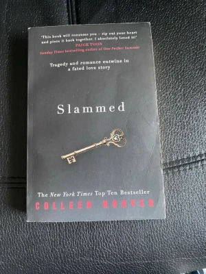 Slammed av Colleen Hoover - Säljer boken 'Slammed' av Colleen Hoover. Omslaget är svart med en nyckel i guld och vit text. Romanen handlar om kärlek, familj och poesi, och är en New York Times bestseller. Perfekt för dig som gillar känslosamma och romantiska berättelser.