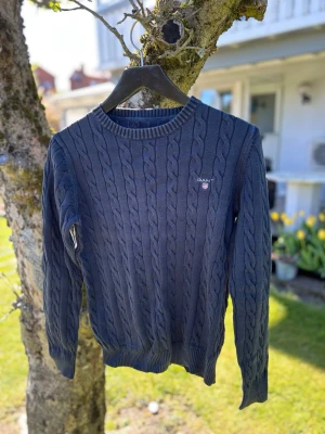 Mörkblå kabelstickad tröja GANT - Kabelstickad mörkblå tröja från GANT med rund halsringning och ribbade muddar. Tröjan har GANT-logga broderad på bröstet och är tillverkad i ett mjukt material som passar perfekt till lager-på-lager-stil.