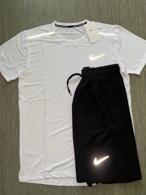 Nike träningsset t-shirt & shorts - Nike träningsset med en vit t-shirt och svarta shorts. Båda plaggen har Nike-logga framtill. T-shirten har rund hals och kort ärm, shortsen har dragsko i midjan. Perfekt för träning eller sportaktiviteter.                                                        ⚠️ STORLEKSINFO: Lappen säger L (Kina), men motsvarar en EU Small (S). • Bröst: 95–101 cm. • Midja: 88–94 cm. Först till kvarn! 🚀 