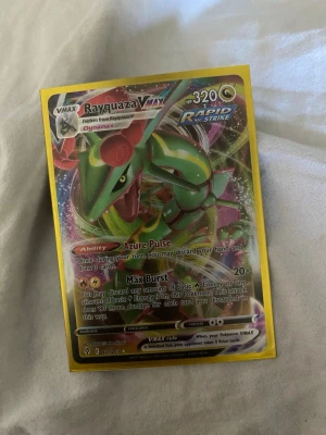 Rayquaza VMAX Pokémonkort - Rayquaza VMAX Rapid Strike Pokémonkort med färgstark design och holografiska detaljer. Kortet har en gul ram och visar Rayquaza i grön, röd och gul färg. Perfekt för samlare eller Pokémon-fans som vill utöka sin kortsamling.