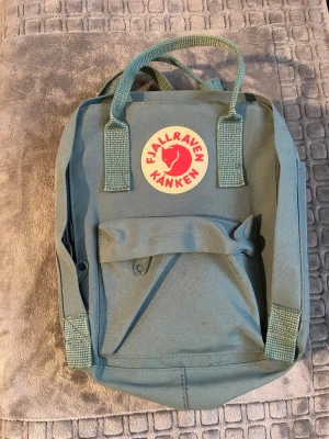 Grön Fjällräven Kånken ryggsäck - Säljer en klassisk grön Fjällräven Kånken ryggsäck med det ikoniska runda röda och vita märket framtill. Ryggsäcken har ett stort huvudfack, en mindre ficka med dragkedja på framsidan och två sidofickor. Tillverkad i slitstarkt syntetmaterial.