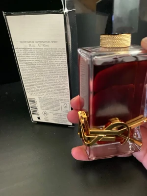 Yves Saint Laurent Libre EdP 90ml - Säljer en lyxig parfym från Yves Saint Laurent, Libre Eau de Parfum, 90ml. Flaskan är rektangulär i glas med mörkröd vätska, svart asymmetrisk kork och guldfärgad metallogo runt flaskan. Perfekt för dig som gillar eleganta och moderna dofter.