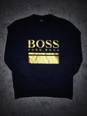 Svart tröja med guldtryck Hugo Boss - Svart tröja från Hugo Boss med stort guldfärgat logotryck på bröstet. Klassisk rund halsringning och långa ärmar. Tröjan har en clean och modern look med metallic-detaljer som verkligen sticker ut.