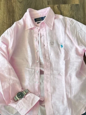 Ljusrosa linneskjorta från Polo Ralph Lauren - Klassisk ljusrosa skjorta från Polo Ralph Lauren med blå broderad logga på bröstet. Skjortan har lång ärm, knappar framtill och en avslappnad passform. Perfekt till jeans eller chinos för en clean look.