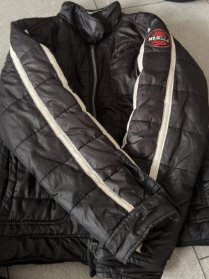Svart pufferjacka från Magilla - Svart pufferjacka från Magilla med vita ränder längs ärmarna och ett broderat märke på axeln. Jackan har hög krage, dragkedja framtill och quiltad design. Perfekt för kalla dagar och ger en sportig vibe.