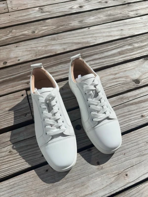 Vita sneakers från Christian Louboutin - Stilrena vita sneakers från Christian Louboutin med klassisk låg profil och ikonisk röd sula. Skorna är tillverkade i slätt läder och har vita skosnören samt diskreta sömmar. Perfekta för dig som vill ha en clean och exklusiv look.