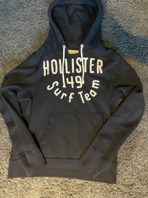 Mörkblå hoodie från Hollister - Mörkblå hoodie från Hollister med vit broderad text 'Hollister 49 Surf Team' på bröstet. Klassisk känguruficka framtill och vita dragsnören i huvan. Perfekt för en avslappnad och cool stil.