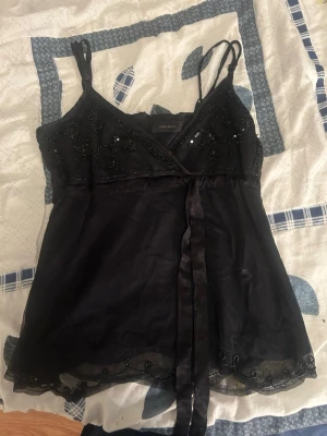 Svart pärlbroderad linnetopp Vero Moda - Supersnygg svart linnetopp från Vero Moda med tunna axelband, pärlbroderier och omlottdetalj framtill. Toppen har ett tunt, skirt material och en dekorativ knytband under bysten. Perfekt för dig som vill ha något extra till outfiten.