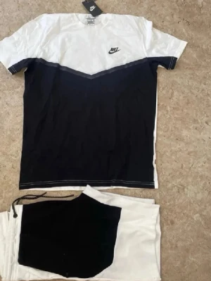 Nike svartvitt set med t-shirt & shorts - Nike set med t-shirt och shorts i svart och vitt. T-shirten har en svart V-formad färgblockning och Nike-logga på bröstet. Shortsen är vita med svart panel och dragsko i midjan. Perfekt för en sportig och avslappnad stil.