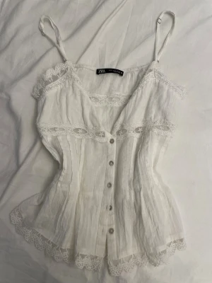 Vit spetsdetaljerad linnetopp Zara - Superfin vit linnetopp från Zara med spetsdetaljer längs kanterna och knappar framtill. Tunt, luftigt material och smala axelband. Perfekt för varma dagar och ger en skön vibe till din outfit.
