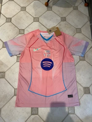 Rosa FC Barcelona matchtröja Nike - Snygg rosa FC Barcelona matchtröja från Nike med klubbmärke och Spotify-logga på bröstet. Tröjan har korta ärmar, blå detaljer vid krage och sidor samt röd och blå grafik. Tillverkad i lätt och ventilerande funktionsmaterial, perfekt för fotbollsträning.