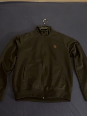 Svart Fred Perry hoodie - Snygg svart hoodie från Fred Perry med dragkedja framtill och två fickor. Ikonisk Fred Perry-logga broderad på bröstet och mönstrade band med loggor längs ärmarna. Hög krage och ribbade muddar. Perfekt för en sportig och stilren look.