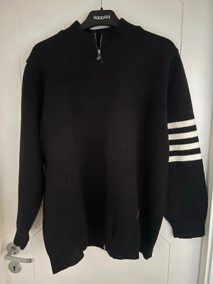 Svart stickad tröja med vita ränder - Svart stickad tröja från Thom Browne med hel dragkedja och tre vita ränder på ena ärmen. Tröjan har en avslappnad passform och ribbade muddar. Perfekt för dig som gillar sportig och enkel stil.