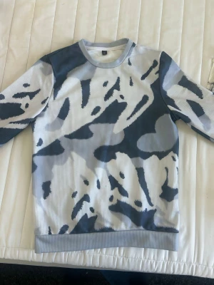 Grå och vit camo-mönstrad tröja - Säljer en långärmad tröja med grått och vitt camo-mönster. Tröjan har rund halsringning och ribbade muddar vid ärmslut och nederkant. Perfekt för dig som gillar streetwear och vill sticka ut med en cool look.