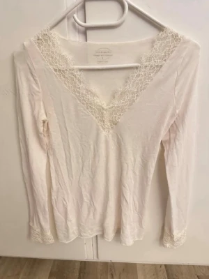 Vit långärmad topp från Intimissimi - Säljer en vit långärmad topp från Intimissimi med v-ringning och spetsdetaljer vid halsringning och ärmslut. Tillverkad i ett ultralätt material med kashmir för extra mjuk känsla. Perfekt för dig som gillar stilrena och feminina plagg.