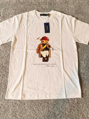 Ralph lauren bear T-shirt Ny-oanvänd (S) - Säljer en helt ny och oanvänd Ralph Lauren Bear T-shirt i storlek M. Stilren vit T-shirt med den ikoniska Polo Bear-designen på framsidan som ger en klassisk och exklusiv look. Tillverkad i mjukt och bekvämt material med hög kvalitet – perfekt för både vardag och streetwear-stil. 🔹 Märke: Ralph Lauren 🔹 Modell: Bear T-shirt 🔹 Färg: Vit 🔹 Storlek: S 🔹 Skick: Helt ny / oanvänd 🔹 Passform: Klassisk Nypris är högt – perfekt möjlighet att göra ett fynd. Hör gärna av dig vid frågor eller funderingar 