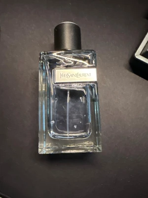 Yves Saint Laurent parfymflaska - Säljer en stilren parfym från Yves Saint Laurent. Flaskan är rektangulär i genomskinligt glas med en svart, räfflad kork och en metallisk etikett med loggan på framsidan. Perfekt för dig som gillar exklusiva dofter och snygg design.