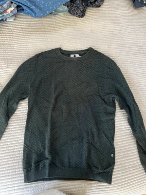 görn tröja - Mörkgrön stickad tröja med diskret strukturmönster och rund hals. Tröjan har långa ärmar och ribbade muddar vid ärmslut och nederkant. Perfekt för dig som gillar en enkel men ändå stilren look.