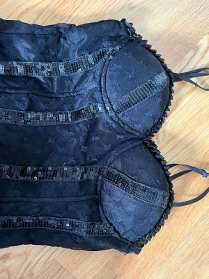 Svart spetskorsett med paljettdetaljer - Säljer en svart korsett med vadderade kupor, spetsmönster och dekorativa paljettränder framtill. Axelbanden är smala och kanten har en söt volang. Perfekt för dig som vill ha något extra snyggt. Använd 1 gång 