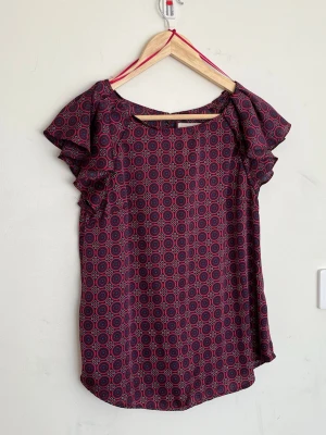 Mönstrad blus med volangärm - Snygg blus i mörkblått och rött med grafiskt cirkelmönster. Blusen har korta volangärmar och rund halsringning. Tillverkad i ett lätt och mjukt tyg som faller fint. Perfekt för dig som gillar färg och mönster.