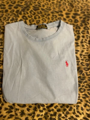Ljusblå t-shirt från Polo Ralph Lauren - Snygg ljusblå t-shirt från Polo Ralph Lauren med klassisk röd logga broderad på bröstet. T-shirten har rund halsringning och korta ärmar. Tillverkad i mjuk bomull som känns skön mot huden. Perfekt för en clean och stilren look.