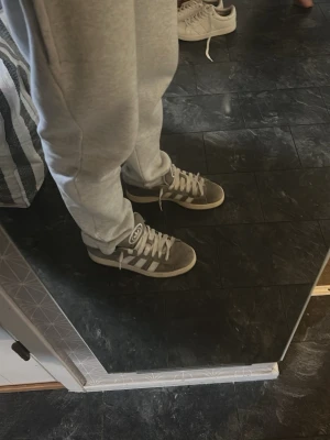 Grå Adidas Campus 00s - Säljer ett par gråa Addidas Campus 00s. Skorna har en klassisk låg modell med bred plös och tjocka vita skosnören. Materialet är mocka och textil, vilket ger en skön och avslappnad vibe. Använda genom en sesong men i bra sick flrutom slitage inuti skon vid hälen. Priset kan förhandlas.😊
