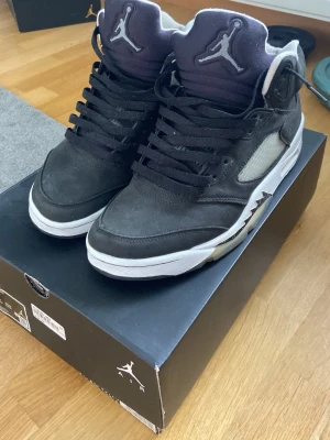 Svarta Air Jordan 5 Retro sneakers - Säljer ett par svarta Air Jordan 5 Retro sneakers med vita detaljer och Jumpman-logga på både plös och häl. Skorna har högt skaft, svarta skosnören och meshpaneler på sidorna. Ovandelen är i mocka och sulan är vit med svarta och grå inslag.