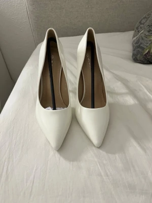 Vita pumps/klackeskor - Eleganta vita pumps i blank finish med spetsig tå och smal stilettklack. Perfekta till bröllop, fest, student, bal eller andra högtidliga tillfällen. Klassisk och tidlös modell som passar lika bra till klänning som till kostymbyxor.  Storlek: 38 Färg: Vit Material: Syntet med blank/lackad yta Spetsig tå Hög, smal klack i fint skick  Säljes då de inte kommer till användning. Hör gärna av dig vid frågor eller om du vill ha fler bilder