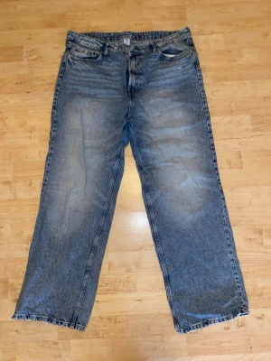 Baggy blå jeans med råa benslut - Baggy jeans i klassisk blå denim med ljusa slitningar och råa, fransiga benslut. Modellen har fem fickor och rak passform. Perfekta för en avslappnad streetstil.