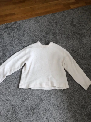 Vit stickad tröja Gina - Mysig vit stickad tröja med rund hals och avslappnad passform. Tröjan har långa ärmar och ribbade muddar vid hals, ärmslut och nederkant. Perfekt för lager på lager och enkel att matcha med olika outfits.