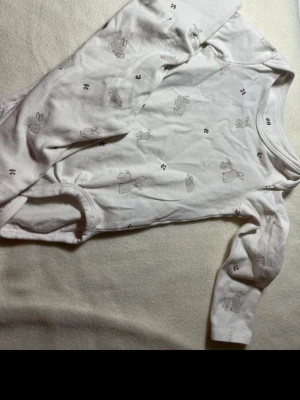 Vit långärmad body med kaniner H&M - Söt vit långärmad body från H&M med gulligt kaninmönster och små blommor. Tillverkad i mjuk bomull, perfekt för mysiga dagar. Tryckknappar i grenen för enkel påklädning. Passar perfekt till de minsta.