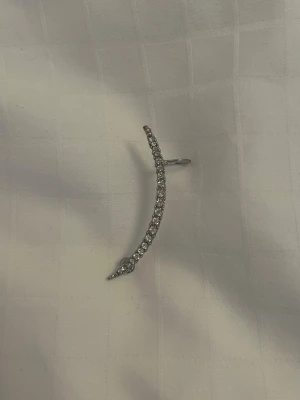 Silvrigt ear cuff örhänge med stenar - Ear cuff örhänge i silverfärg med en böjd form som följer örats kant. Örhänget är dekorerat med gnistrande små stenar längs hela framsidan. 