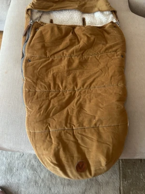 Brun åkpåse med foder - Mysig brun åkpåse från Voksi i manchestertyg med mjukt vitt foder på insidan. Åkpåsen har dragkedja på sidan och ett brunt lädermärke med två fjädrar framtill. Perfekt för kalla dagar i vagnen.
