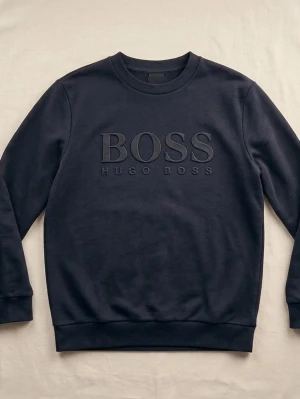 Svart sweatshirt från Hugo Boss - Stilren svart sweatshirt från Hugo Boss med stor BOSS-logga i relief på bröstet. Tröjan har rund halsringning, långa ärmar och ribbade muddar vid ärmslut och nederkant. Tillverkad i mjukt bomullsmaterial för en bekväm känsla.