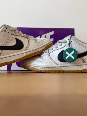 Nike Dunk Low Vit/Svart Gum - Nike Dunk Low sneakers i vitt läder med svart Swoosh och gummifärgad yttersula. Klassisk siluett med lågt skaft och vita skosnören. Perfekt för dig som gillar retrovibbar och streetwear. Kommer med originalkartong och StockX-tag. Pris kan diskuteras.