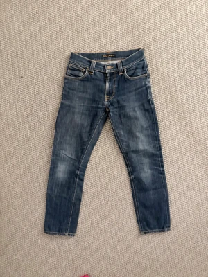 Blå jeans från Nudie Jeans - Snygga blå jeans från Nudie Jeans med klassisk femficksdesign och gul kontrastsöm. Jeansen har en rak passform och är tillverkade i denim med lätt tvättad look. Perfekta för en avslappnad stil. W28 l32