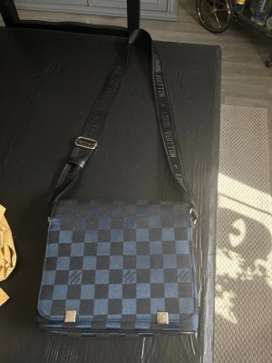  axelväska blå-rutig - Snygg axelväska  i blått och svart damier-mönster. Väskan har justerbar axelrem med logotyp, fyrkantig form och detaljer i metall. Perfekt för dig som vill ha en stilren och exklusiv accessoar till din outfit.